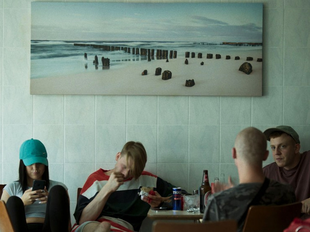 A group of young people share drinks at a table, focused on their phones, below a cheap photo print of a beach