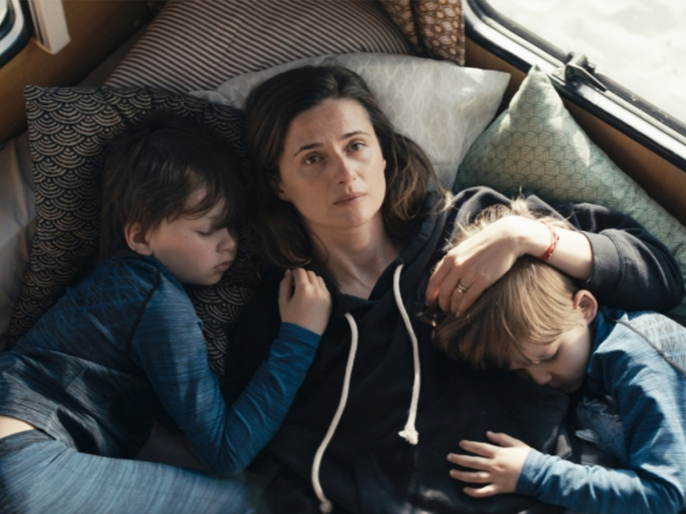 A mother holds her two sleeping children, resting on a caravan bed as she looks up at the ceiling, tired