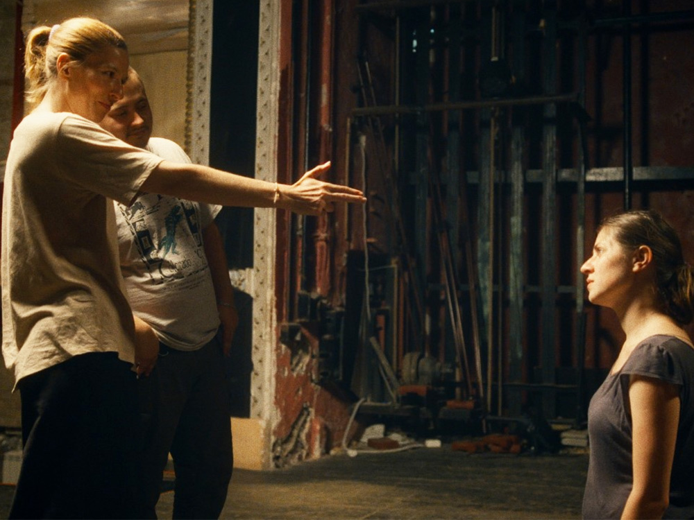 At a theatre backstage, an actor mimics a gun with her fingers at a kneeling actor. Another actor casually observes