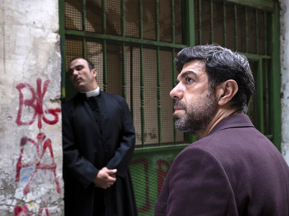 Two priests stand by a barred window in a graffittied street of Naples