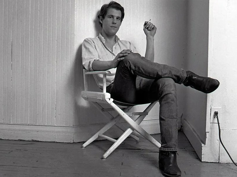 Artist Edward Brezinski sitting on a fold up chair in a house, one leg up on the other, in a casual white shirt, jeans, smoking a cigarette. The photo is black and white.