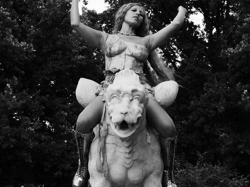 Lafawndah sits atop a fountain statue of a mythical beast, arms splayed out.