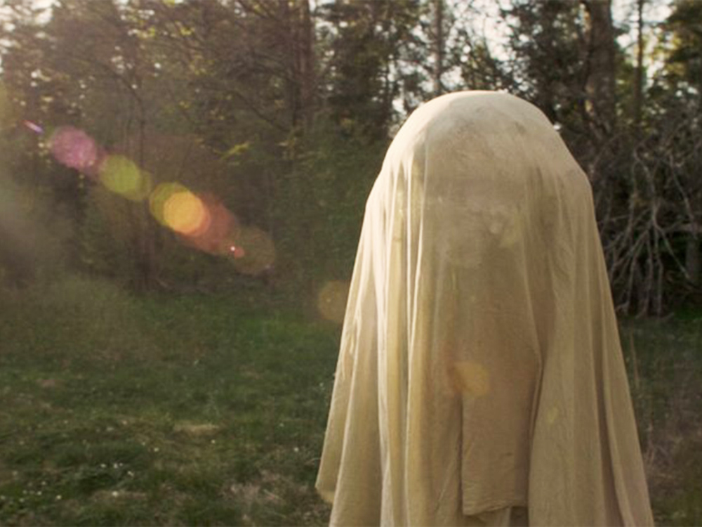 A young person hides under a white cloth while standing outside in the sunlight, in a Swedish forest