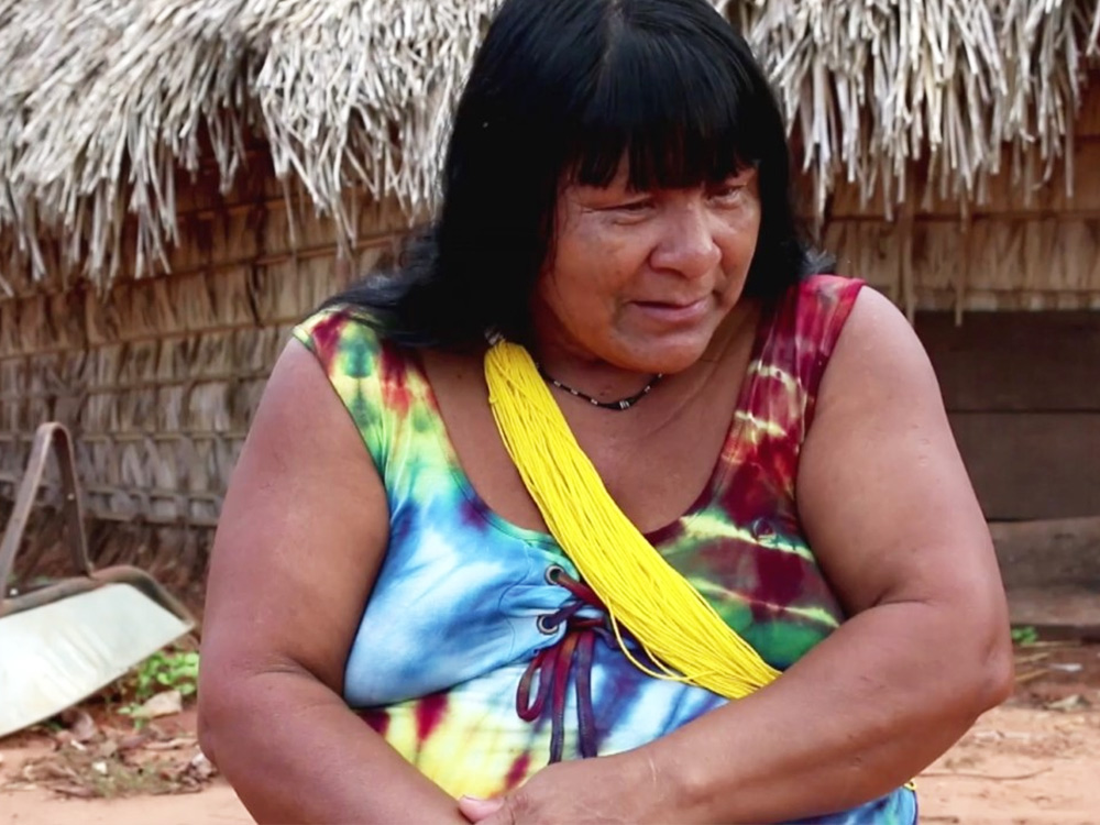 An Ikpeng woman dressed in colourful clothes sits in front of their home, staring off