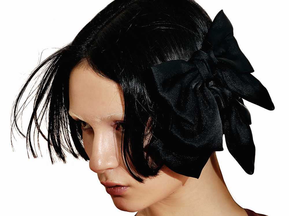 Side profile of musician Evita Manji against a bright white background, wearing a black velvet bow in their hair