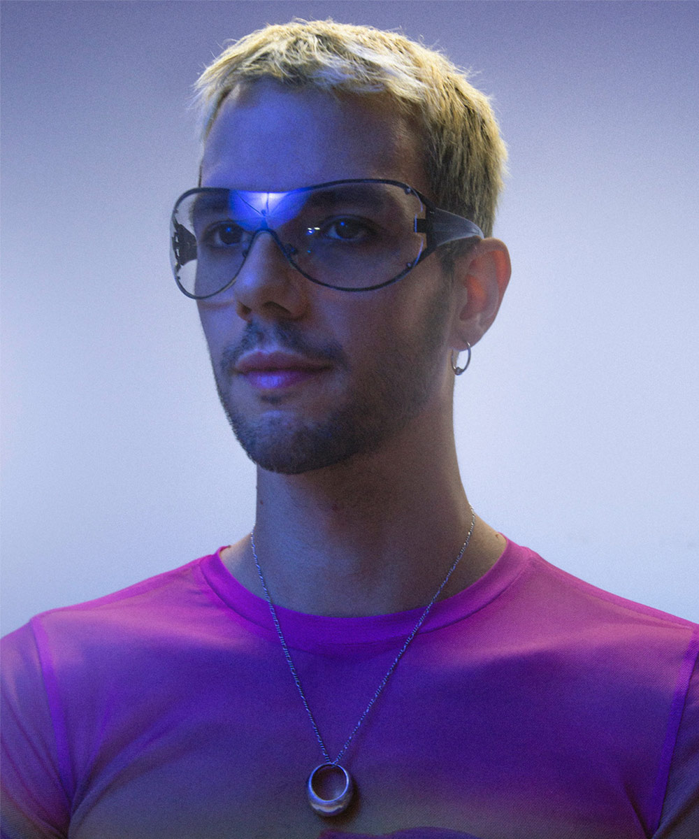 marum is lit by light purple light, cropped blonde hair, bulbous clear sunglasses, and a ring hanging from their neck