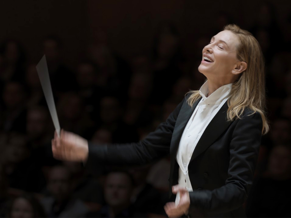 In a suit, Lydia Tár waves a baton at an orchestra offscreen, eyes closed in rapture