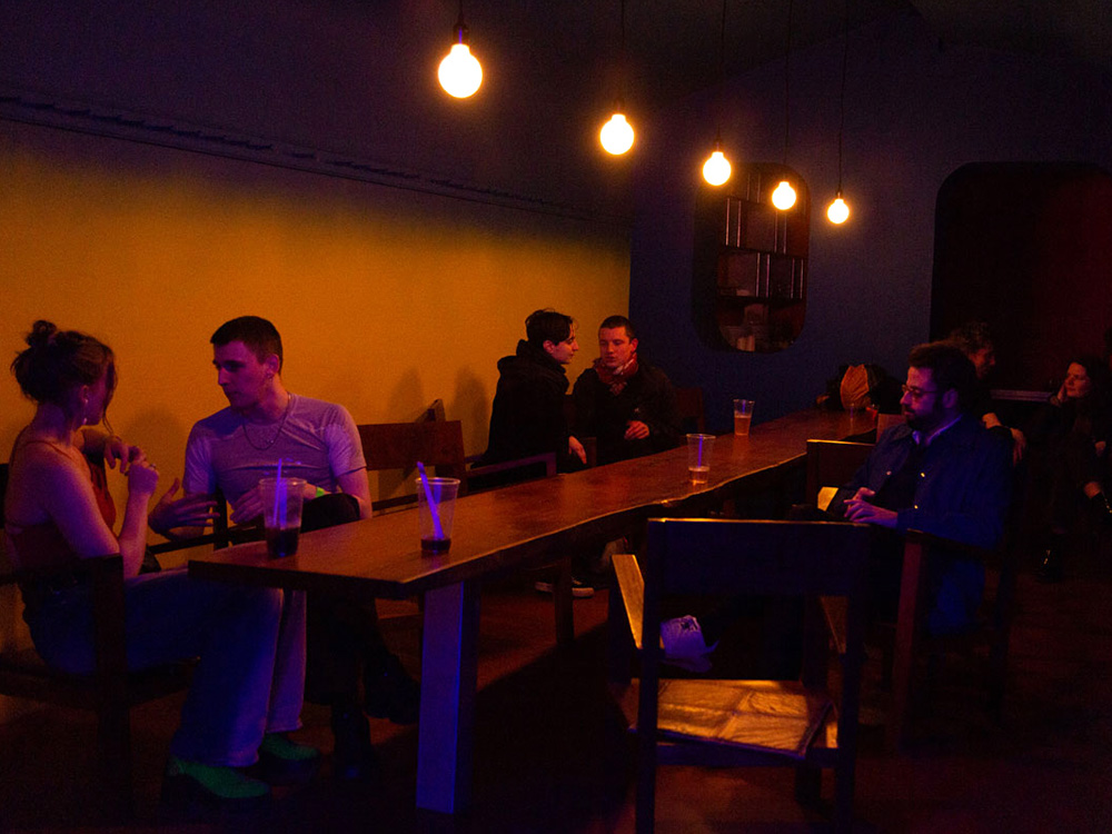 A group of people sit in the Sake Bar, a long wooden table in moody yellow and red lighting