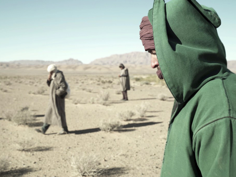 A person in a green hooded jumper walks through the desert, with others in the distance doing the same