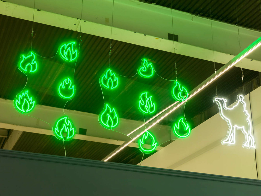 A cluster of green neon flames and a white neon camel hang above the exhibition space