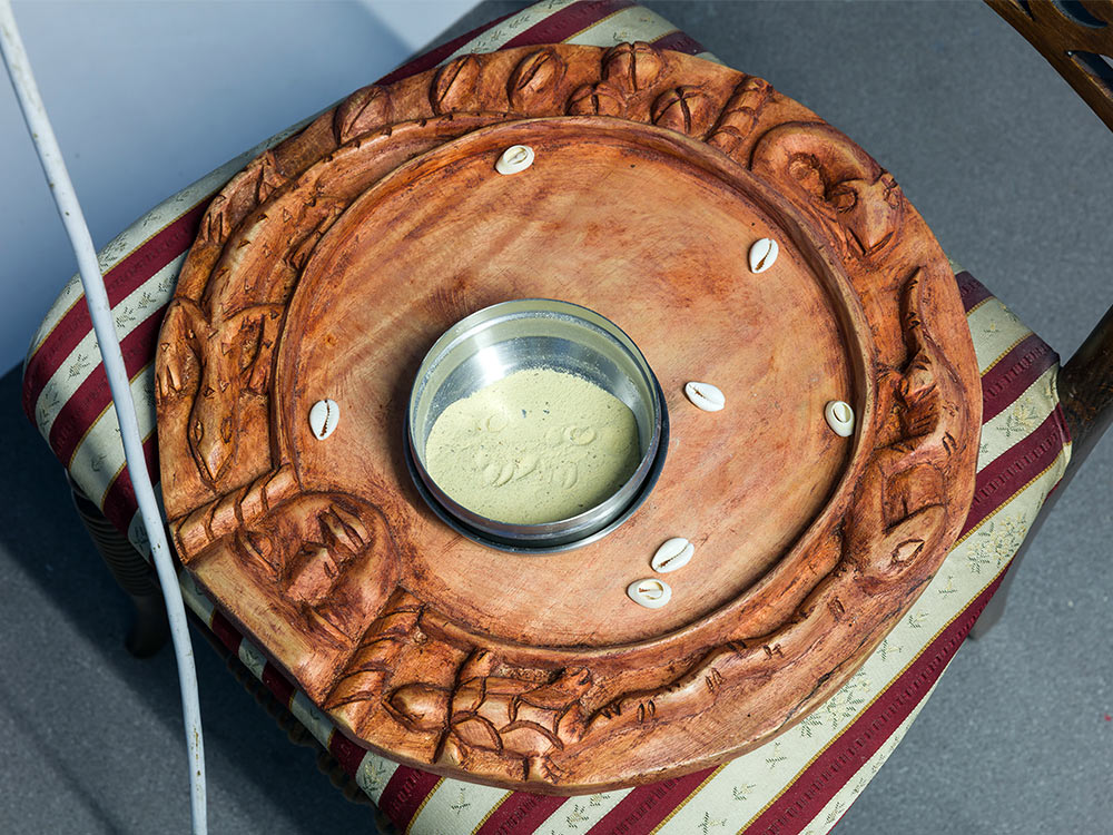 A wooden bowl sitting on a chair. In the bowl is sand and cowrie shells