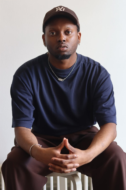 Portrait of Nicolas-Tyrell Scott, sitting on a bench, hands poised. He is Black, wearing maroon pants, a navy short-sleeved top, earrings, and a NYC cap