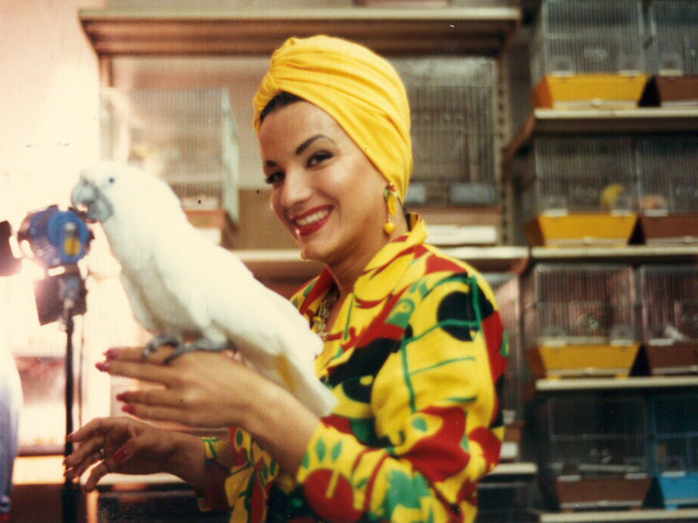 Actress Carmen Miranda is dressed in bright clothing, smiling, white parrot on her arm