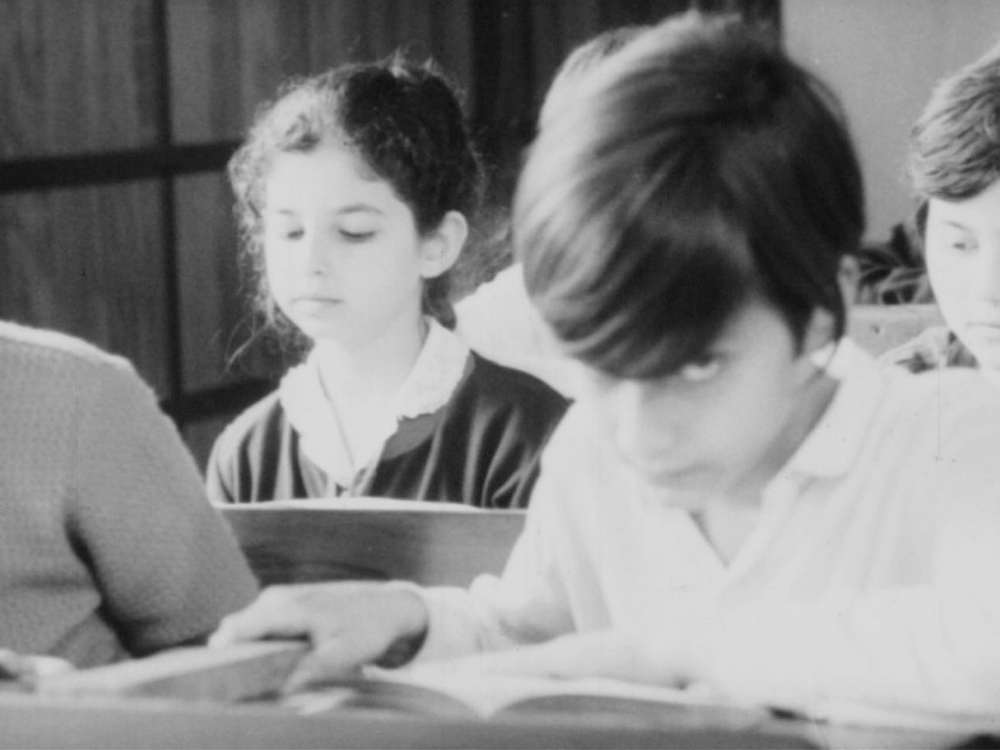 Two young people sit in a classroom. One is looking down at their desk. The other is looking up from a book at the camera, eyes hidden behind their fringe