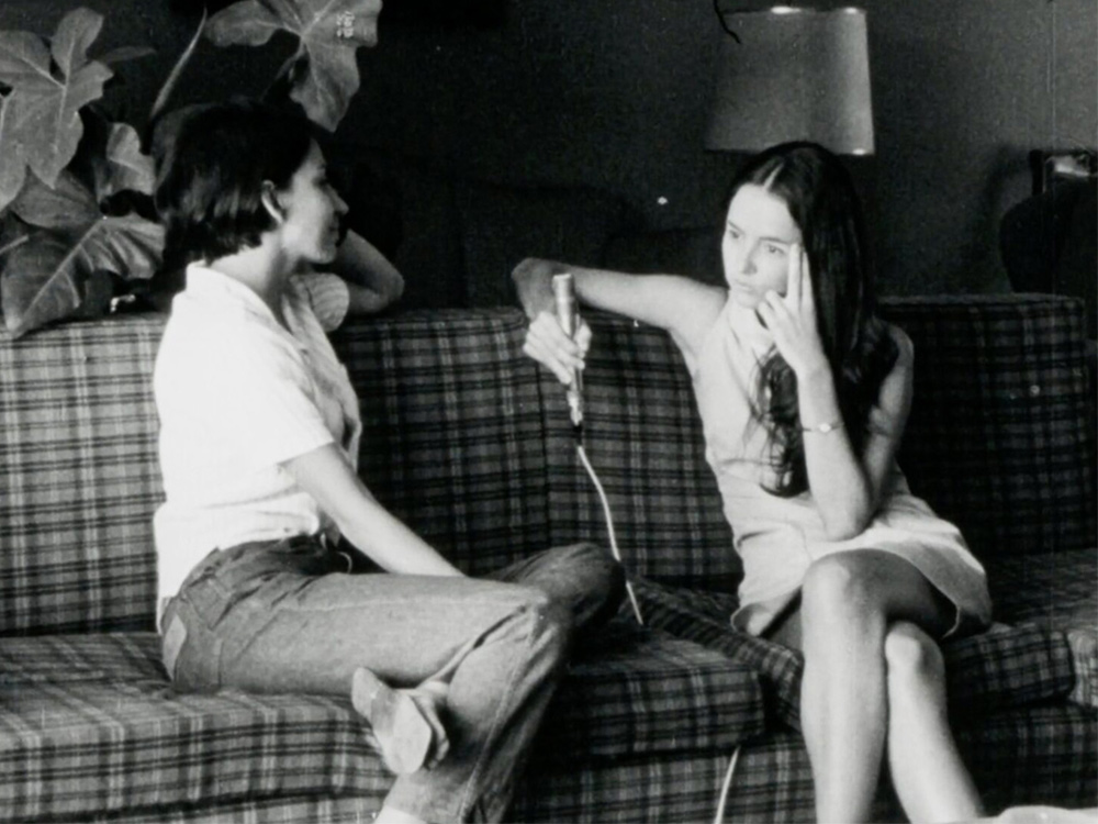 Two young women sit on a couch holding a microphone, thinking about their lives