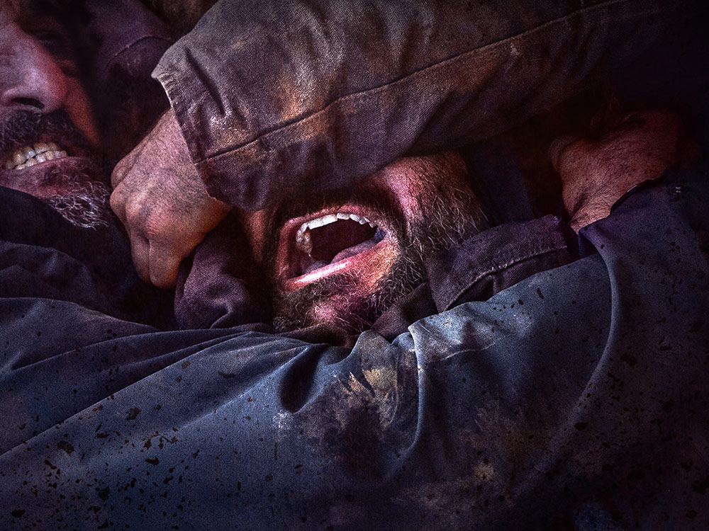 A bearded man shouts with open mouth as his head is held tightly by the arms of other people