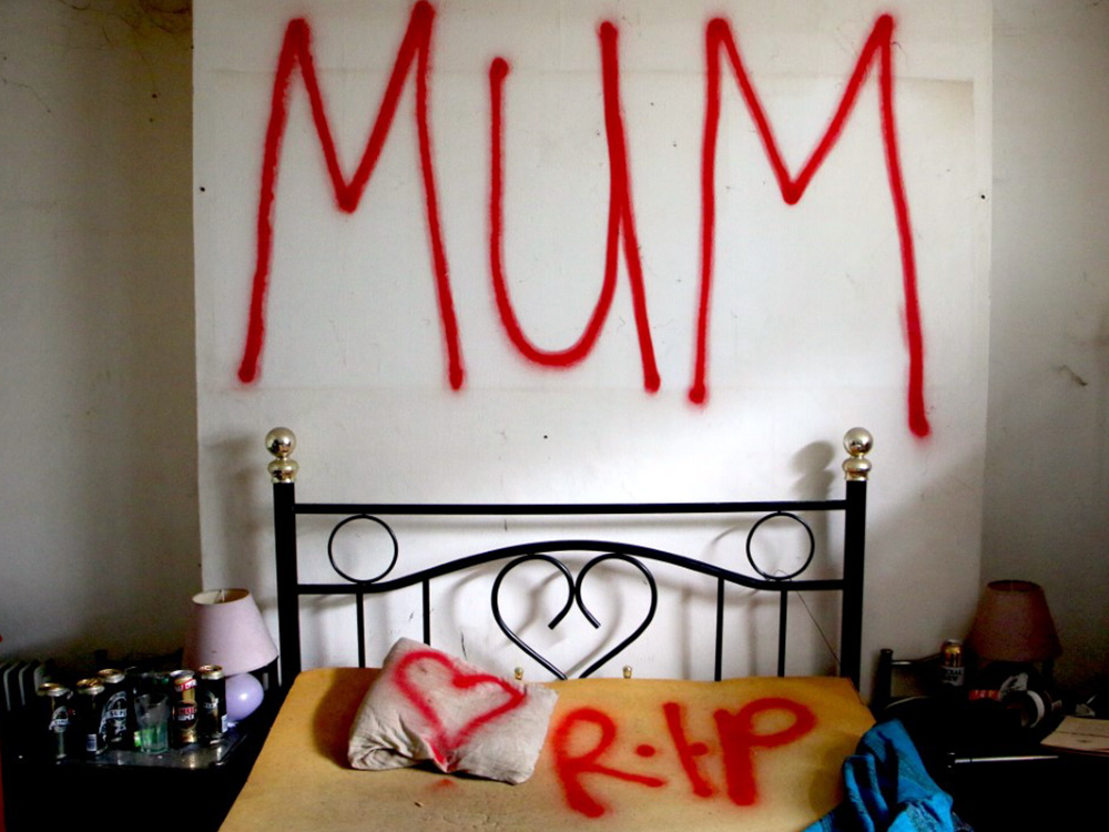 A bed in a house, in red spray paint 'MUM' is written on the wall above, a heart sprayed onto the pillow, and the letters 'R.I.P.'