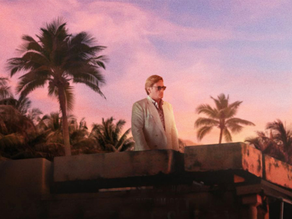 A white man in a white suit and sunglasses stands on a rooftop against palm trees and a pink sunset