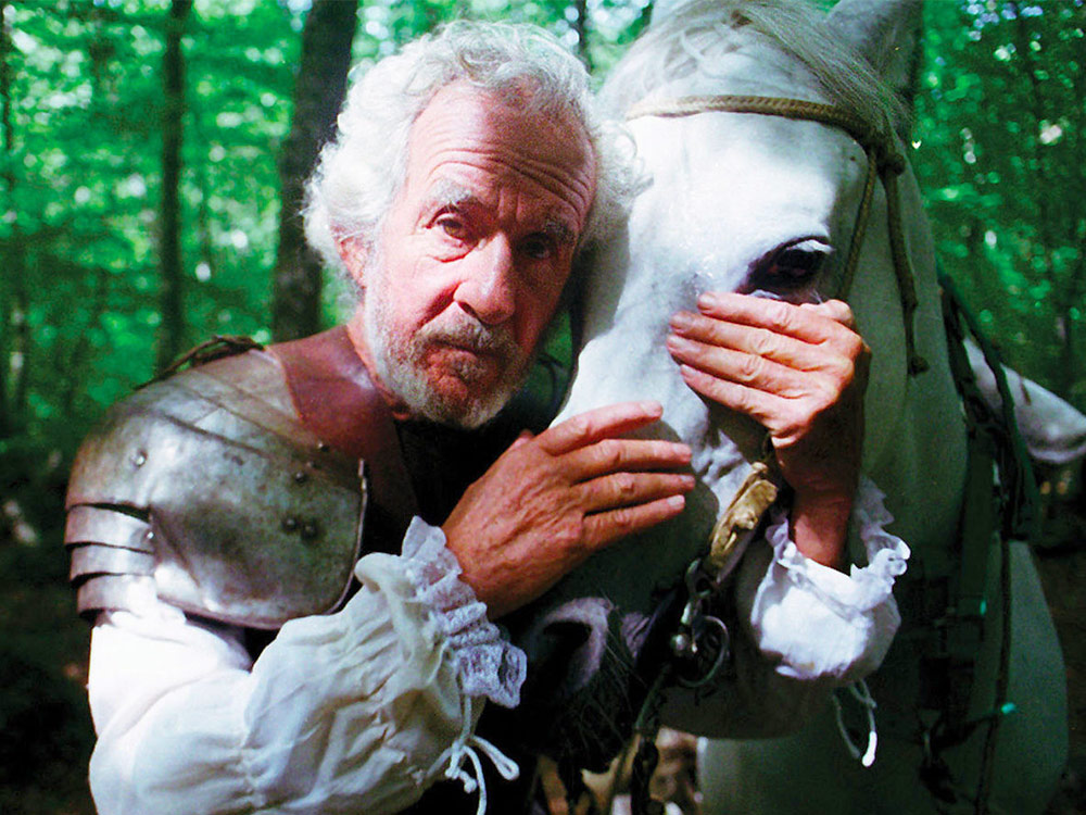 An elderly Don Quixote in knight's armour stares at the camera while he embraces the head of his white horse