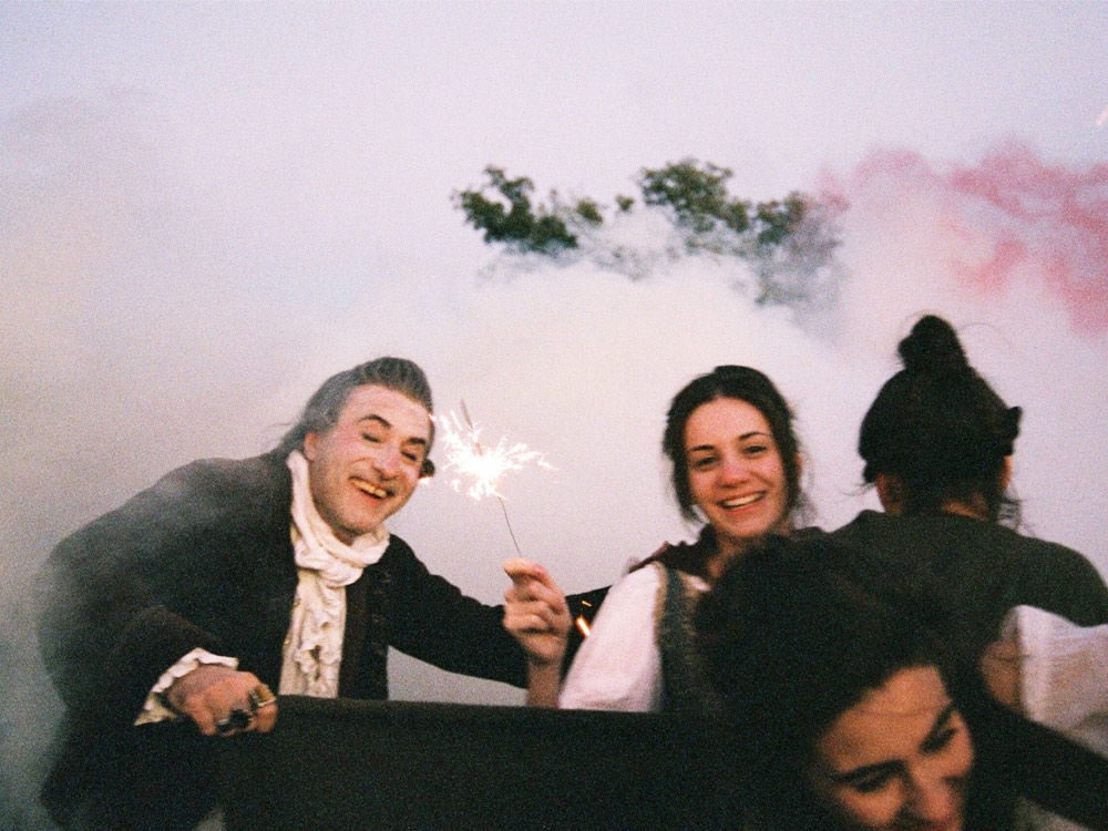 Two people in 18th century European dress wave a sparkler among a joyful group of people, against a wall of smoke