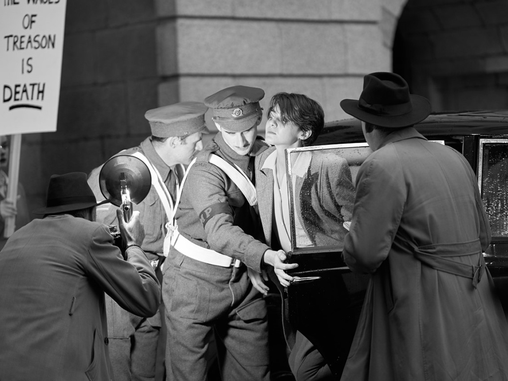 A short-haired person is led out of a car by police. They are being photographed by the media.