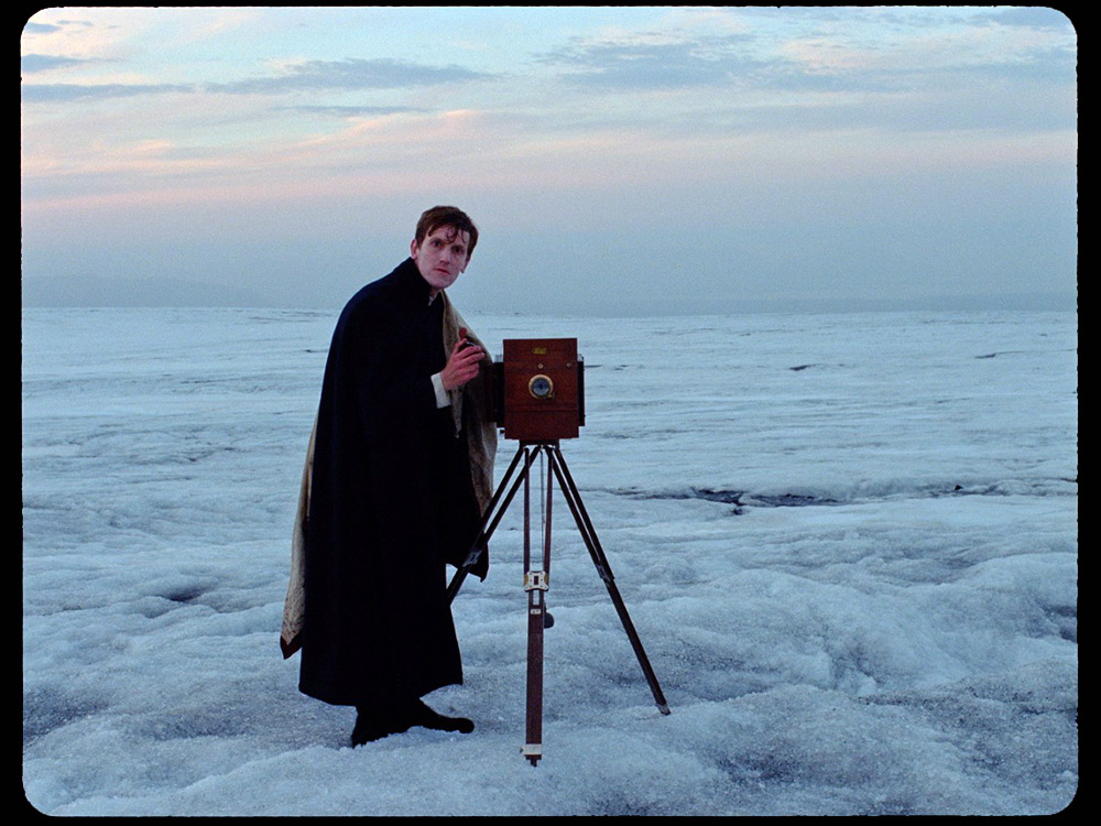 A white man stares at the camera, with an Academy camera, on the snow
