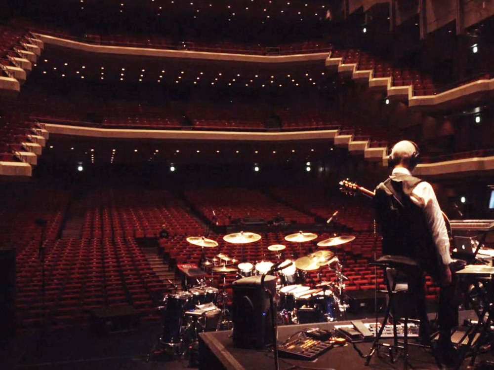 Musician Robert Fripp sits on stage, holding his guitar, staring out at an empty concert hall. There's a lone drum kit on the stage