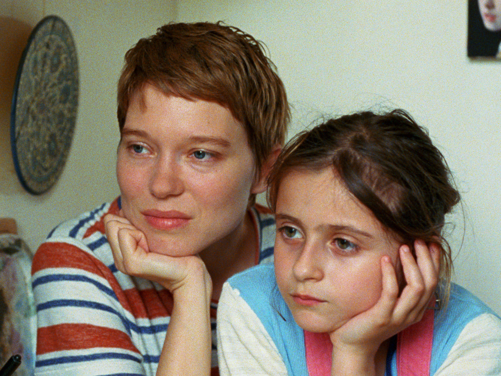 A mother and her daughter sit by each other in their home, hands against faces, staring off camera