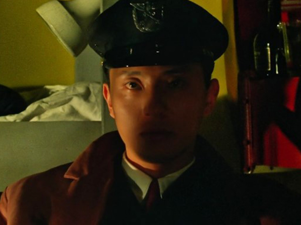 A blank-faced train guard in uniform stares past the camera, half his face in shadows