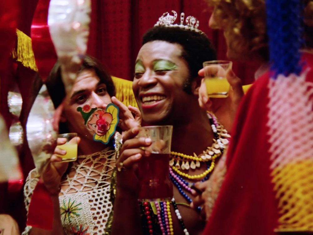 A black person with green eyeshadow, wearing a tiara and covered in beans, surrounded by friends enjoying a drink at a party