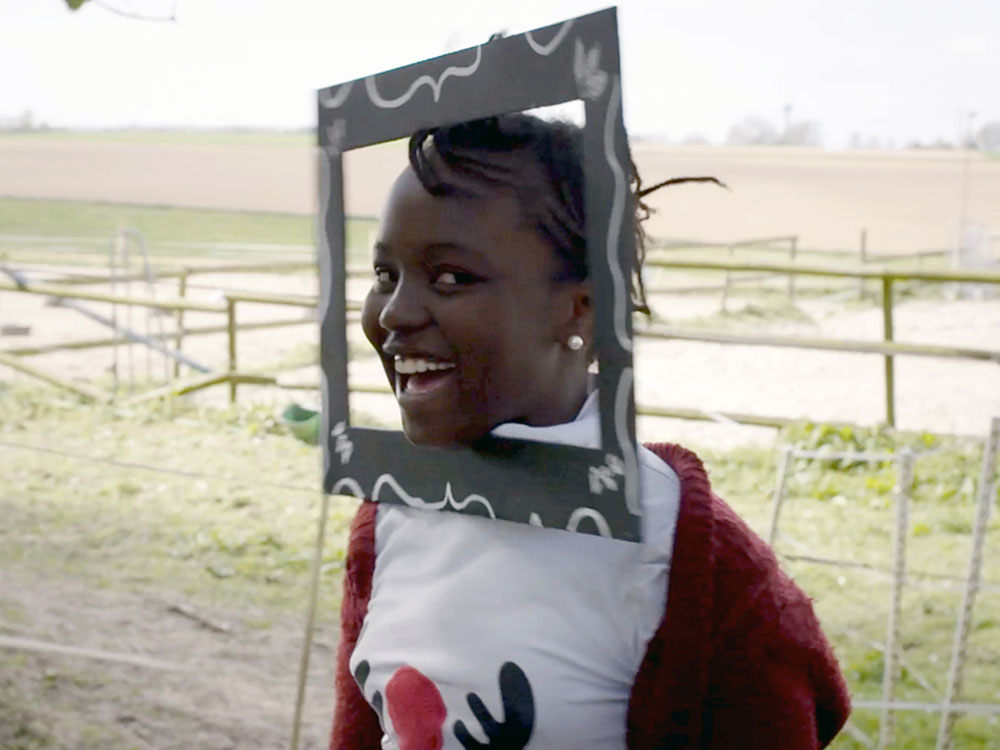 A black child sticks their head through a photo frame they made, smiling widely