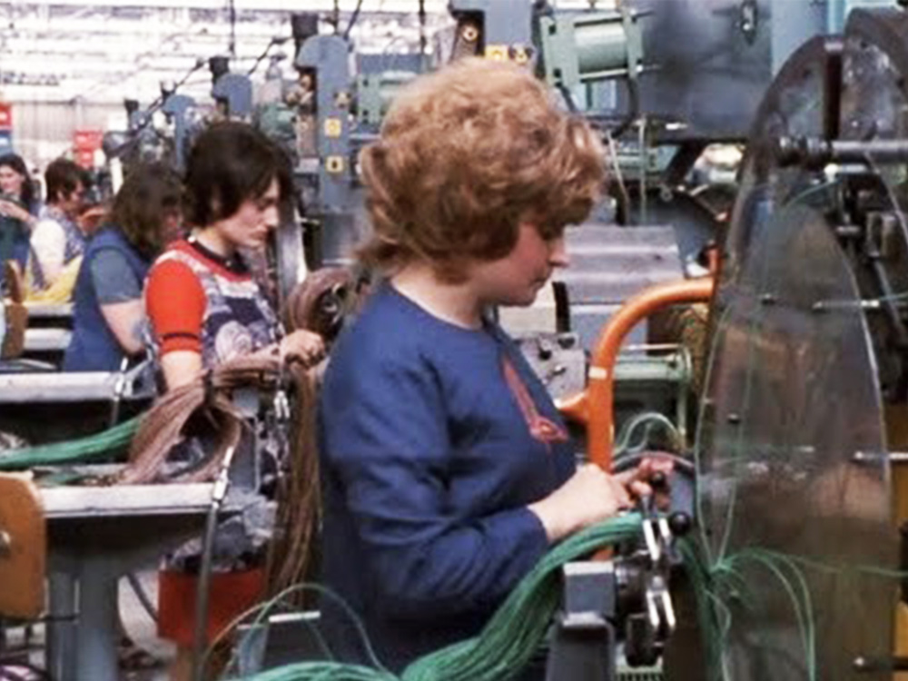 A woman dressed in 70s clothing works in a car factory