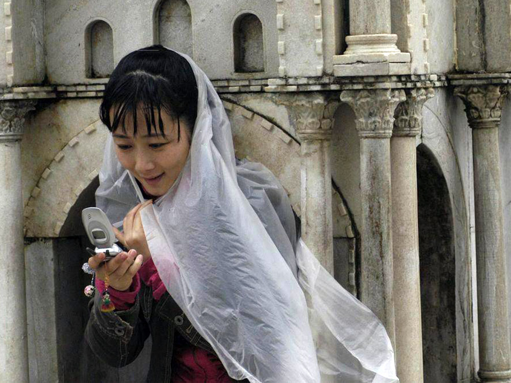 A Chinese woman takes shelter under a plastic poncho and checks her fliphone. She's standing in front of a replica of an ancient building