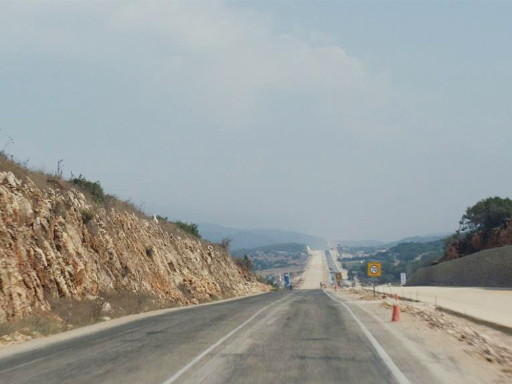 A long road next to a low cliff, stretching into distant rocky hills. The sky is a murky grey-blue