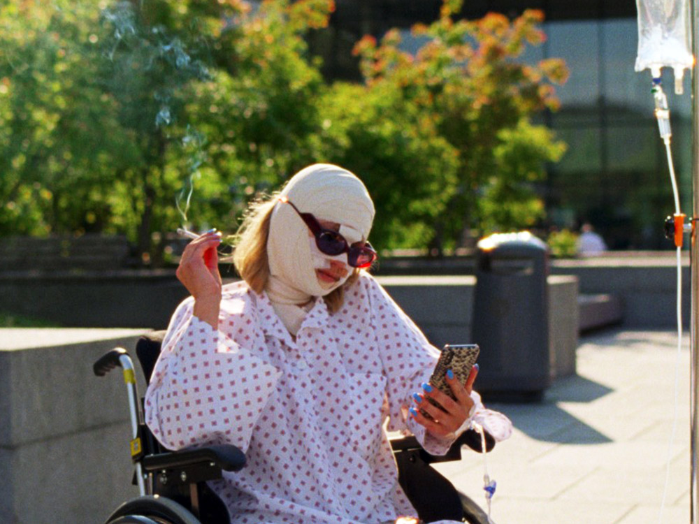 A person in a wheelchair, their face is covered by bandages and sunglasses. They're smoking a cigarette and checking their phone. They have blue nails.