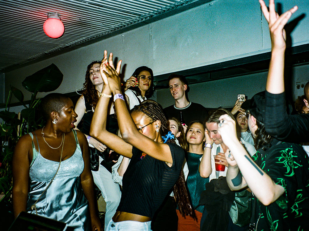 A crowd dances in the Upper Bar of the ICA, hands raised, under a single red globe light