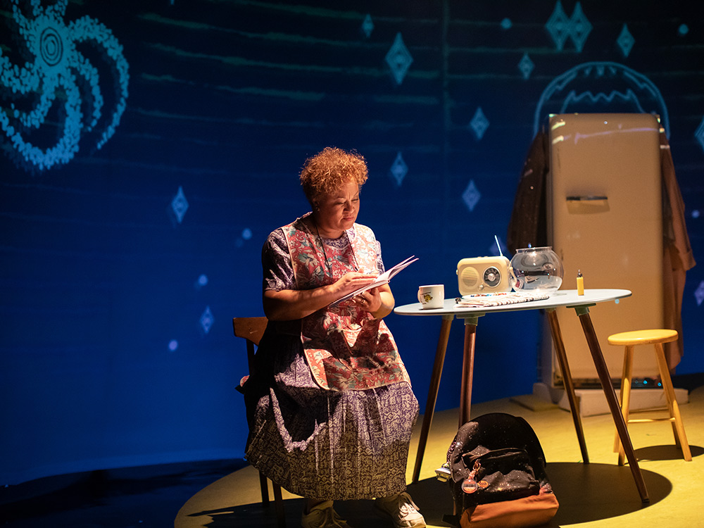 MRS sits on a chair reading the newspaper. On the dinner table is an old style radio and a fishbowl. Behind her is a fridge. There are animated stars and galaxies on a dark blue backdrop