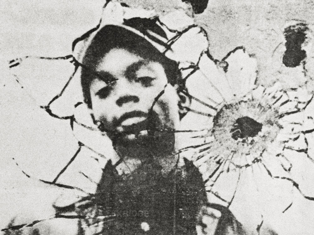 The face of a black man, in black and white, reflected in shattered glass with a bullet hole