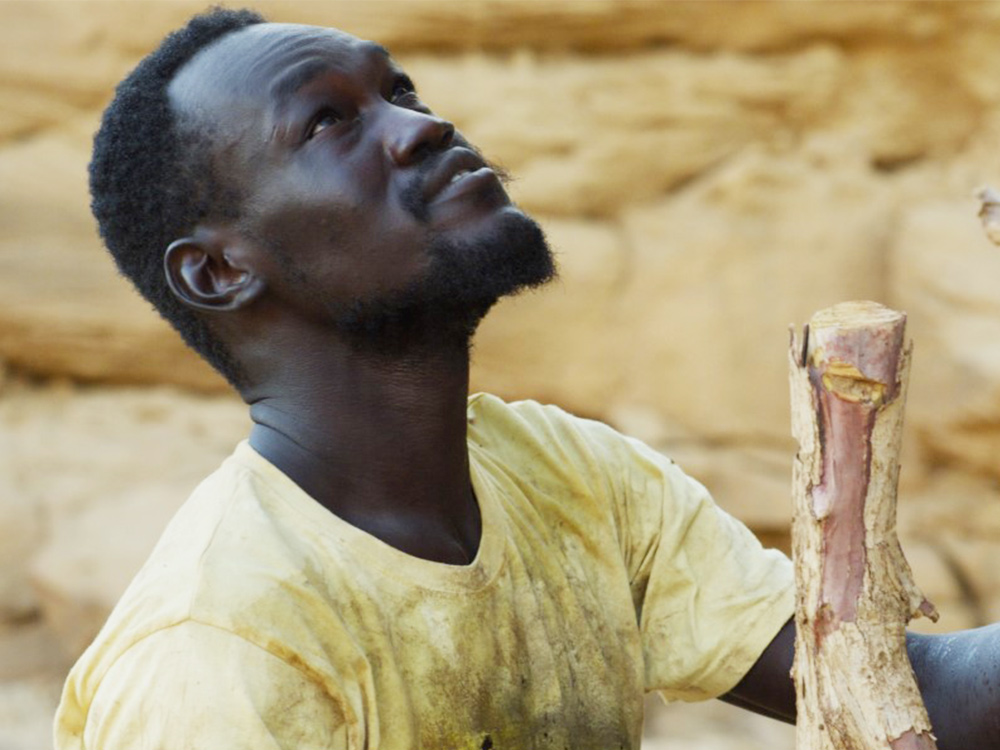 A black man working on the construction of a dam looks up at something tall out of the frame. He's holding a large stick