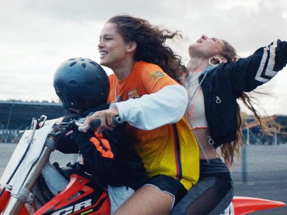 Two people with long hair ride a motorbike fast. The passenger has their arms spread out