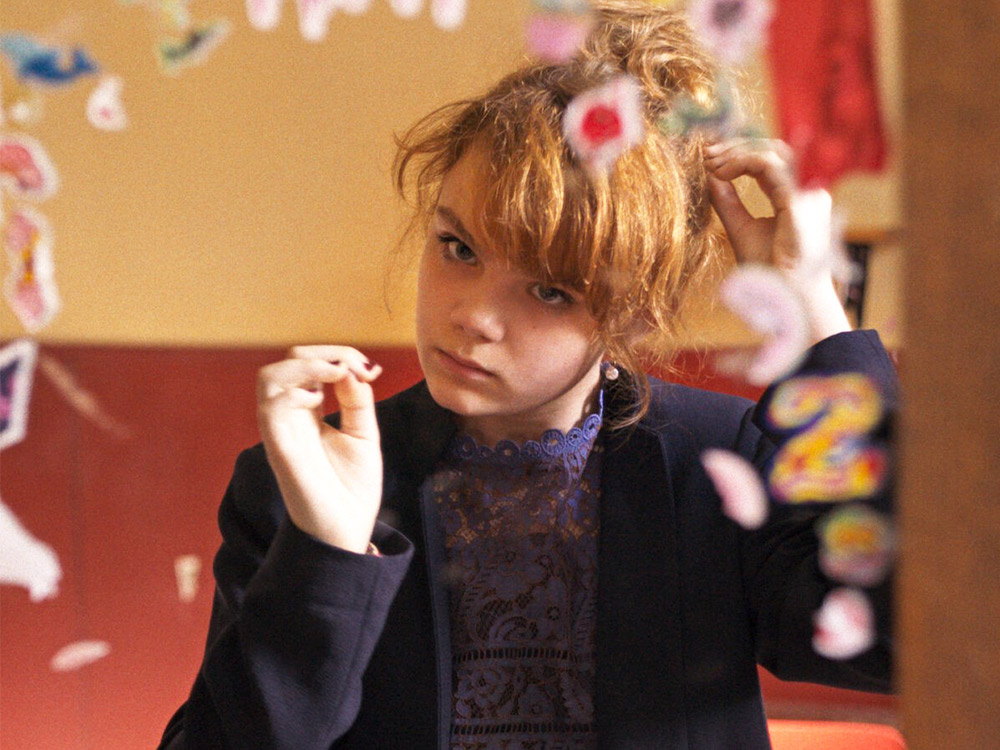 A young woman looks at herself in a child's mirror, dressed in formal clothes and doing her hair