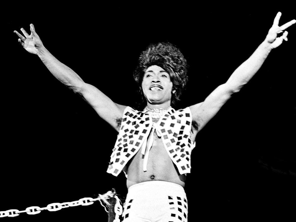 In black and white, Little Richard stands on stage with arms outstretched, peace signs