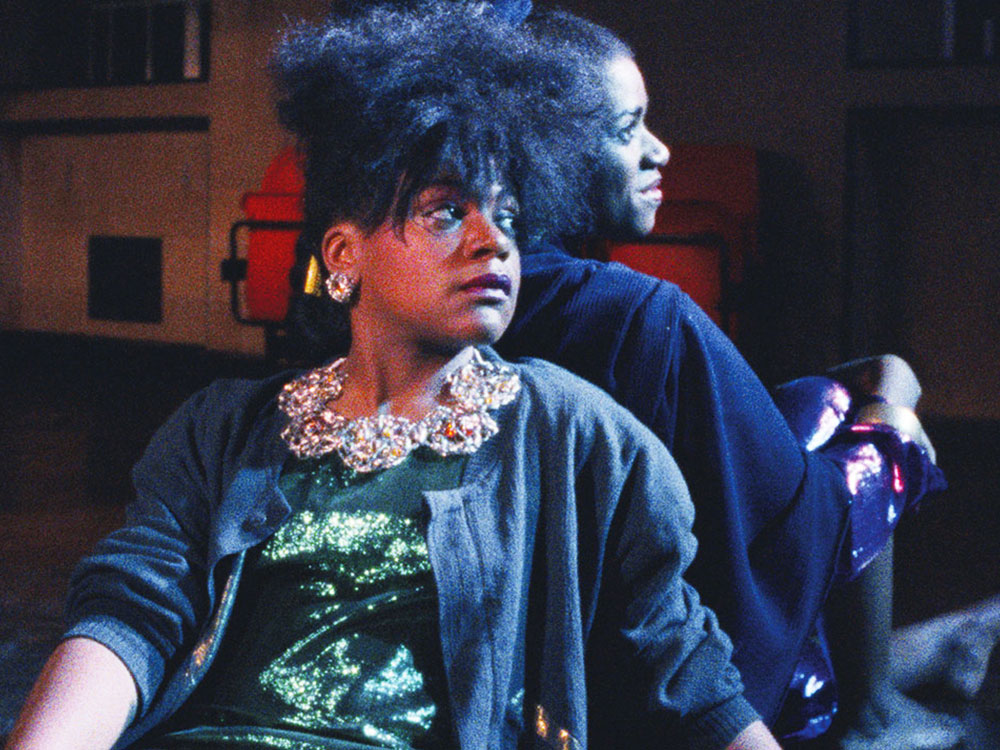 Two black women sit with their backs against one another, looking out