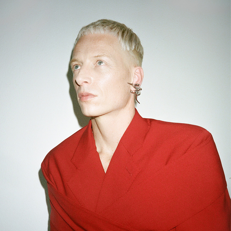 A headshot of Bendik Giske, a white person with short blonde hair, sharp earrings, and red clothing