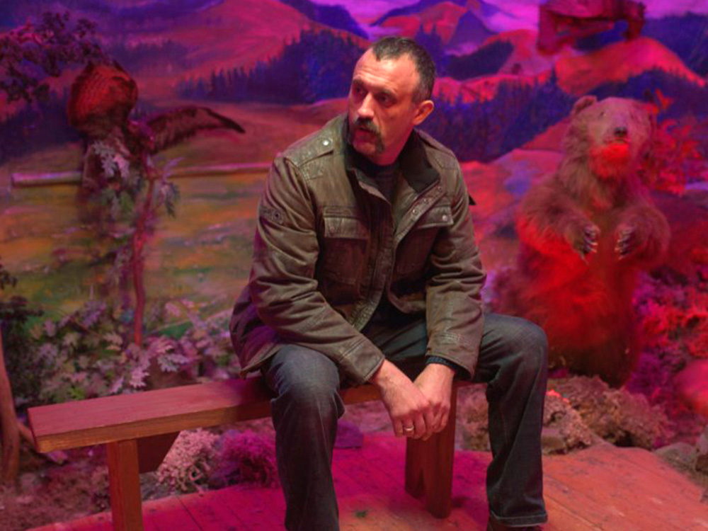 A father sits on a wooden bench under pink light. He looks tired but relieved. Behind him is a model bear and a fake hilly landscape scene