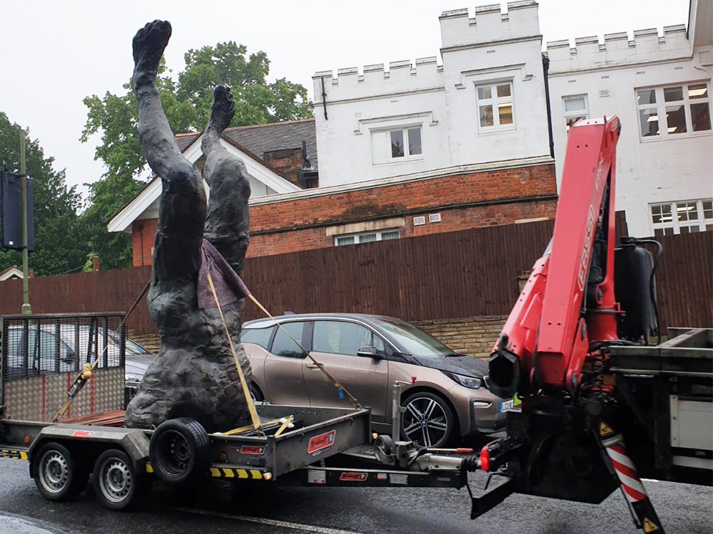 A stone sculpture torso upside down on a tray being driven by a truck, in front of old houses