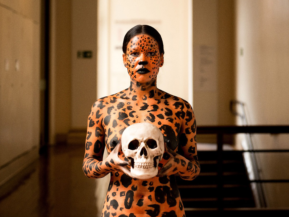 A woman faces the camera, covered in cheetah print orange and black paint, and holding a skull