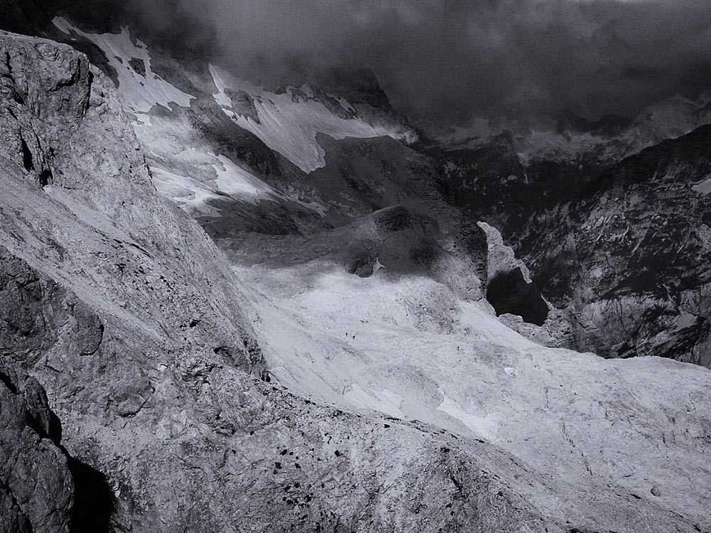 A black and white landscape of a stony mountain range. The shadows create a layer of dark grey over the bright light reflecting off smooth stone