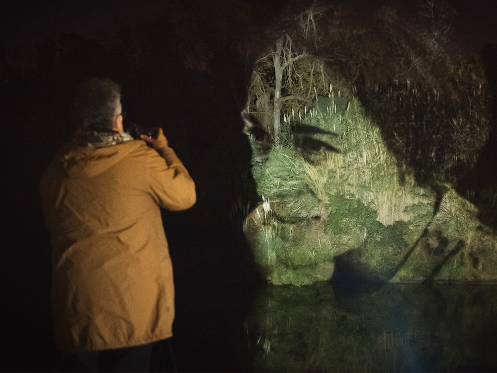 A person takes a photo of a woman's face transposed onto images of grassy ground