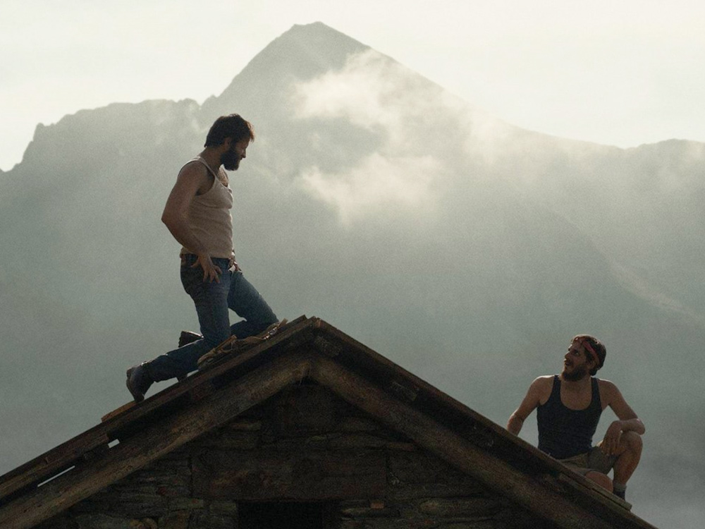 Two bearded people in singlets and jeans sit atop a roof with a cloudy mountain peak in the background